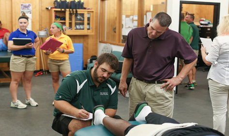 a student sits next to a patient on a bed, with an instructor looking over his shoulder and two other students looking on